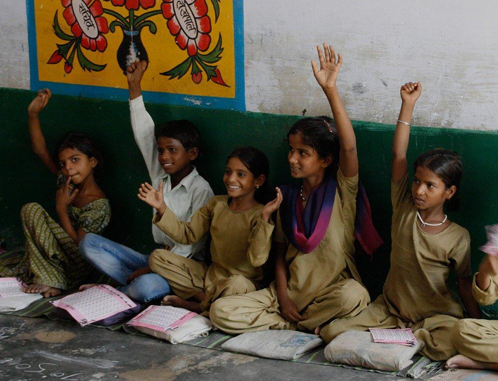 photo of students sitting on the floor of a classroom with hands in the air