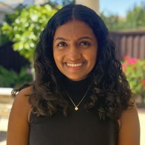 Headshot of an Indian-American woman, 22 years old, with curly black hair, a black tank top, and a necklace. She is smiling, and there is some blurred greenery in the background.