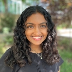 Headshot of an Indian-American woman, 22 years old, with curly black hair, a black shirt, and a necklace. She is smiling, and there is some blurred greenery in the background.